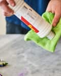 Hands cleaning a marble surface with a green cloth and cleaning solution, surrounded by crayons and a yellow cap.