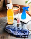Cleaning supplies on a wooden table, including disinfectant spray, hand sanitizer, blue spray bottle, mask, and cloth.