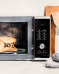 Microwave with open door, steaming paper bag of popcorn inside, surrounded by kitchen utensils.