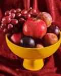 Yellow bowl with pomegranates, plums, and grapes on a red velvet fabric.