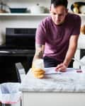 Man in a purple t-shirt standing in kitchen, with trash on the counter next to him. He is writing in a notebook to keep track of what he throws away