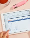 entering expenses into a budget spreadsheet on an ipad with a pink background, calculator, and mug