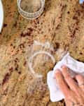 A hand cleaning a granite countertop with a white cloth, a glass, and a white pot nearby.