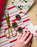 A hand writing a gift tag on a wooden table, surrounded by wrapping paper, scissors, mini wreaths, and decorative ribbon.