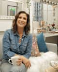 Woman in denim shirt holding a mug, sitting on a gray sofa in a cozy room with blue velvet chair and decorative items.
