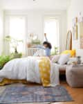 Woman stretching on a bed with white and yellow bedding, surrounded by plants and wall art in a bright bedroom.