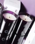 Three lit candles in dark glass holders with white boxes labeled "Beauty Pie" on a purple surface.