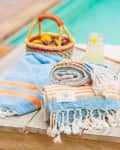 Blue and orange striped towels on a wooden table by a pool, with a basket of fruit and two lemon drinks.