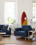 Living room with blue sofa and armchair, surfboard decor, bookshelf, plants, and abstract wall art.