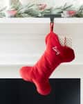 Red bone-shaped stocking with "Lucy" embroidered, hanging on a mantel with festive greenery.