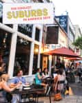 Outdoor dining scene with people at tables under red umbrellas, surrounded by trees and shops, in a lively suburban area.