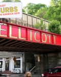 Red railway bridge with "Ellicott City" sign, cars below, and lush green trees in the background.