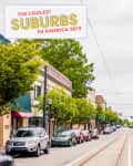 Street view of a vibrant suburban area with parked cars, trees, and colorful storefronts under a cloudy sky.