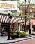 Street view of a charming suburb with brick buildings, striped awnings, and outdoor seating at a café.