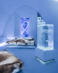 Ice hotel room with fur-covered beds, ice sculpture, and illuminated ice wall art.