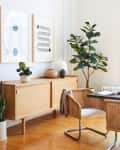 Mid-century modern dining room with wooden sideboard, abstract art, woven chairs, and potted plants.