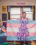 Person holding a trans pride flag with "There Are Trans People Here" text in a room with colorful decor and posters.