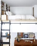 Loft bed with white bedding, bookshelves, and a black ladder above a wooden sideboard with decor and framed art.