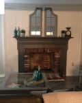 Living room with brick fireplace, dark mantel, decorative bottles, plants, and a glass coffee table.
