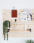 Wooden wall organizer with hanging plants, notebooks, and art prints, next to a pink vase with white flowers.