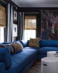 Blue sectional sofa in a living room with eclectic decor, large painting, and antelope head mount on dark walls.