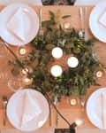 Table set for six with white plates, folded napkins, cutlery, and a centerpiece of greenery and candles.