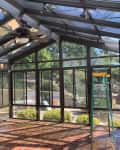 Glass sunroom with terracotta tile floor, green ladder, and ceiling fan, overlooking a garden.
