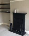 Empty living room corner with black fireplace, beige walls, black shelves, and a gray carpet.