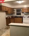 Dated kitchen with wooden cabinets, white stove, red countertops, and a folding chair against tiled wall.