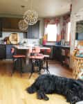 Cozy kitchen with dark cabinets, pink bar stools, floral curtains, and a large black dog on wooden floor.