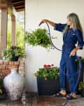 A woman in a denim jumpsuit waters hanging plants on a porch, surrounded by potted flowers and greenery.