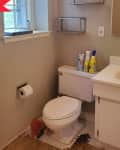 Small bathroom with beige walls, toilet, sink, cleaning products, and a window.