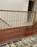 Wooden staircase railing with white spindles on polished hardwood floor, colorful rug in foreground.