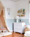 A kids' bedroom with half-walls painted blue, white furniture, and a sand-colored canopy above a reading nook.