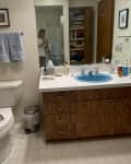 Bathroom before renovation. dated faux wood cabinets, white counter, blue sink, off white walls, white tile floor