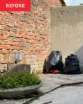 Patio before decorating/renovation. One red brick wall, another plain gray wall, a couple plants, 2 trash bags, no furniture