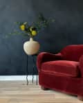 Red velvet armchair next to a ceramic vase with yellow flowers on a black stand, set against a dark wall.