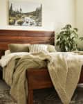 Cozy bedroom with wooden bed, green bedding, knit throw, large plant, and landscape artwork above headboard.