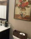 Plant and toiletries atop white sink with dark brown cabinet in bathroom with floral art on beige wall and basket for toilet paper.