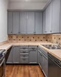 Gray kitchen cabinets with a beige tile backsplash, stainless steel stove, and wooden floor.