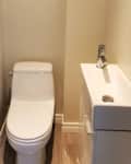 Small bathroom with a white toilet and a compact sink, light-colored walls, and wooden flooring.