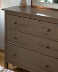 Light gray dresser with three drawers, a white vase, and a mirror on top, next to a white wastebasket.