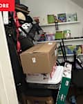 Cluttered storage closet filled with boxes, toys, books, and bags, with a small black table and a chair.