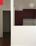 A small kitchen area with maroon cabinets, a white tiled backsplash, and a pendant light, viewed from a living space.