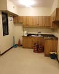 Small kitchen with wooden cabinets, a sink, a water jug, cleaning supplies, and colorful bags on the floor.