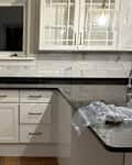 White kitchen cabinets with black granite countertops, marble backsplash, and a pendant light above the sink.