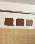 Three brown paint swatches on a white wall above beige tiled shower, with an orange loofah hanging nearby.