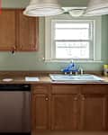 Kitchen with wooden cabinets, stainless steel dishwasher, sink with blue tape, and light fixtures above.