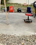 A wooden ladder stands on a concrete surface surrounded by gravel, with a red rolling stool and various buckets nearby.
