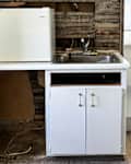 A small kitchen area with a white mini fridge, sink, and open cabinet space, featuring exposed wooden walls and unfinished flooring.